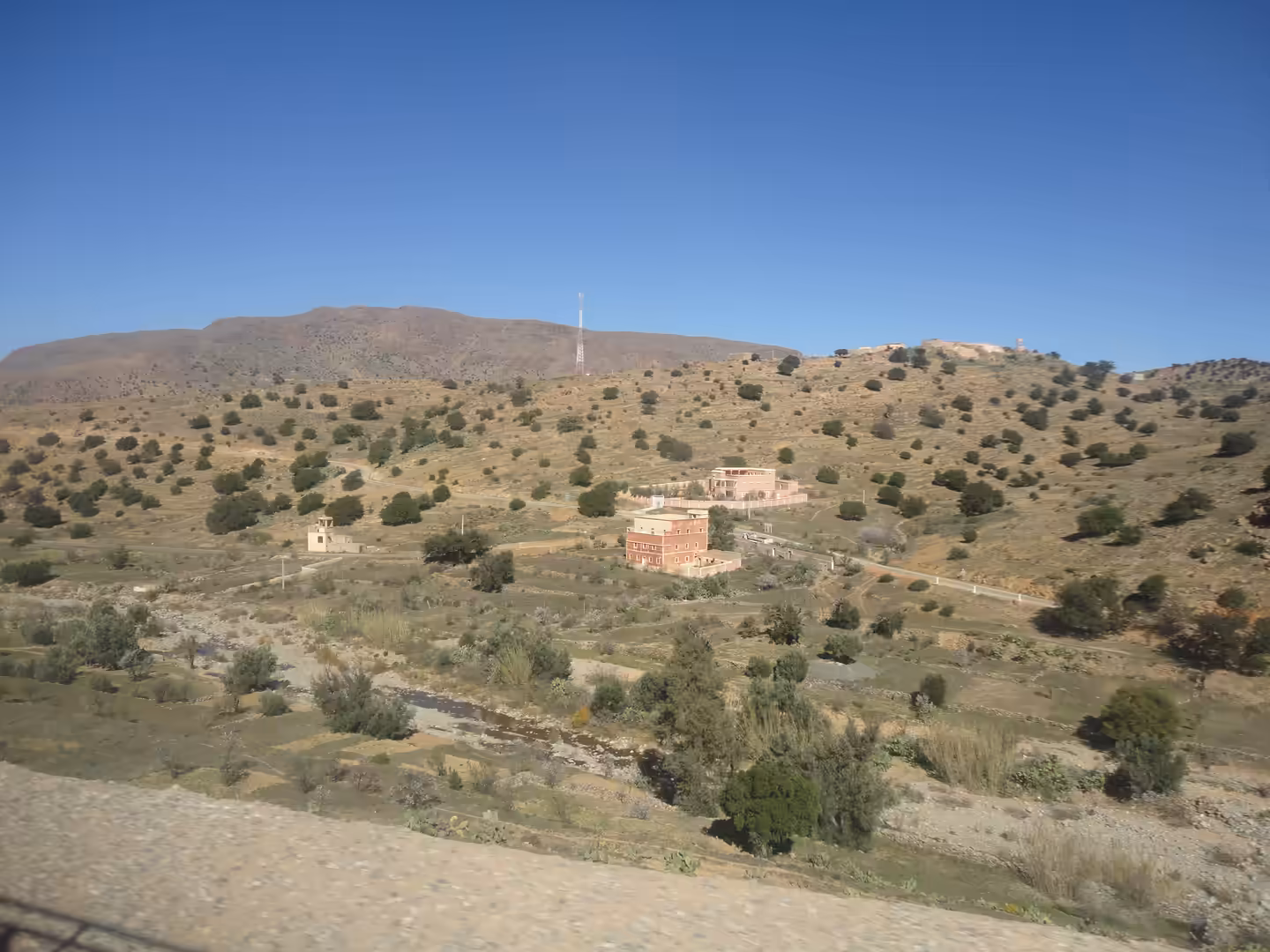 Scattered houses and terraced land in a dry Anti-Atlas valley, with shrubs, a wadi, and rounded hills under a clear sky.