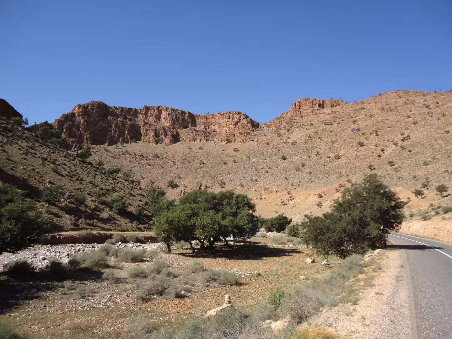 Arid mountain valley with scattered trees, rocky slopes, and a paved road cutting through southern Moroccan highlands.