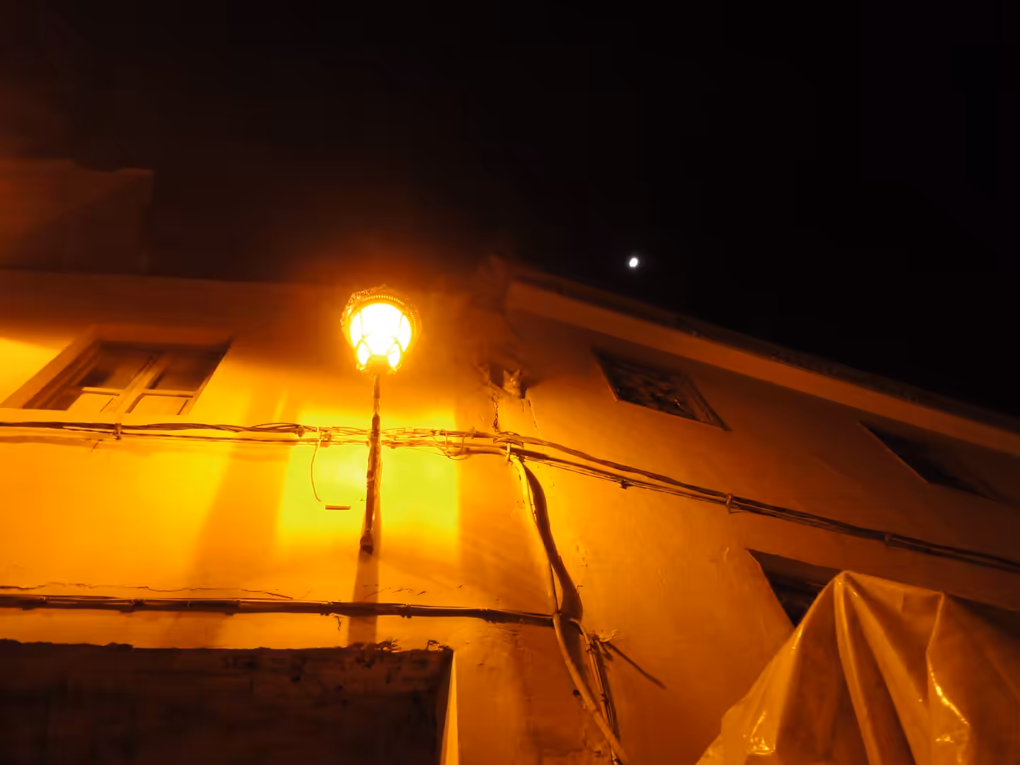 Warm streetlamp illuminating an ochre wall in Taroudant at night, with exposed cables and the moon visible in the dark sky.