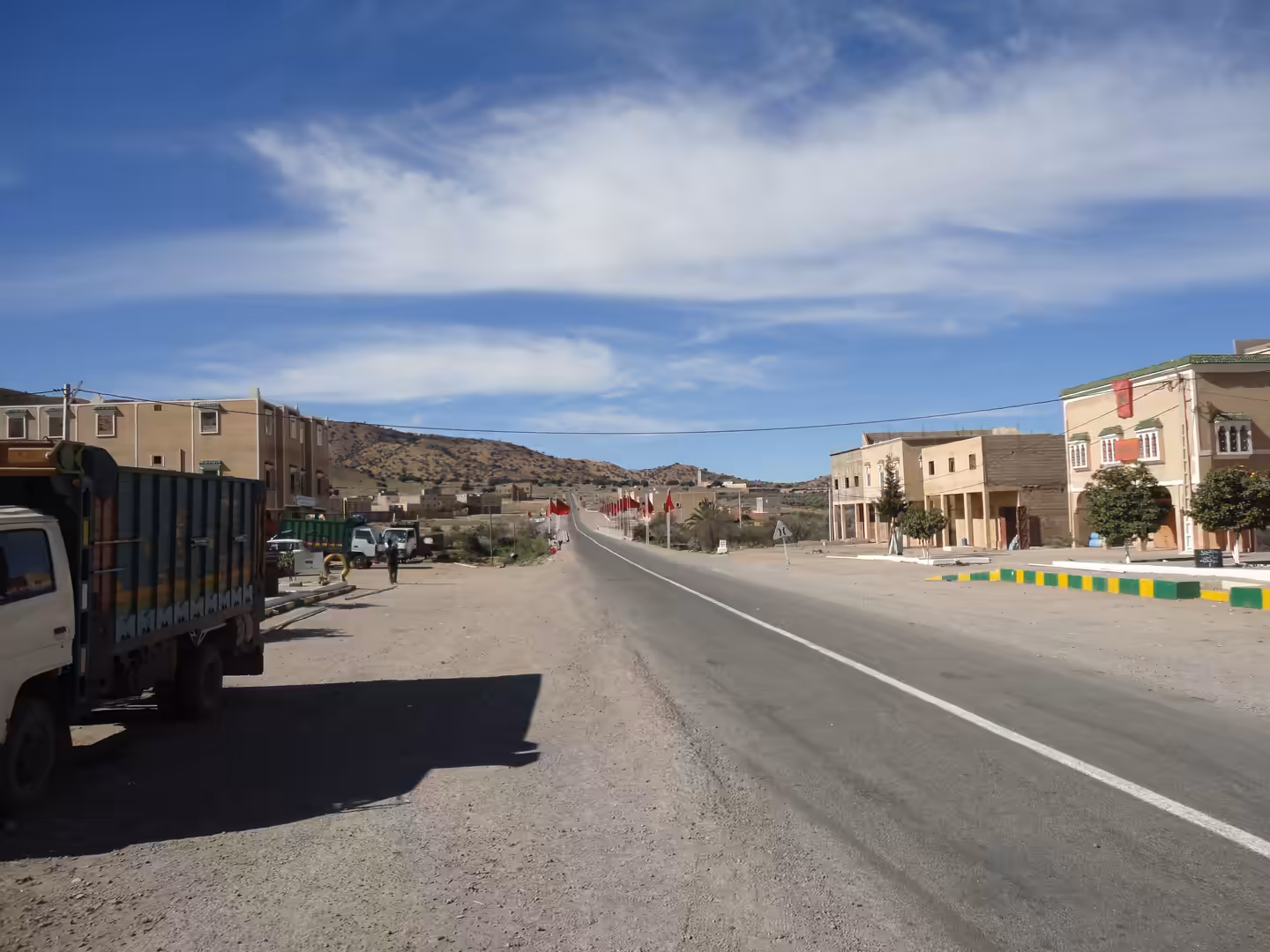 Paved road entering a small district of Ouarzazate, lined with low buildings, parked trucks, Moroccan flags, and arid hills in the background.