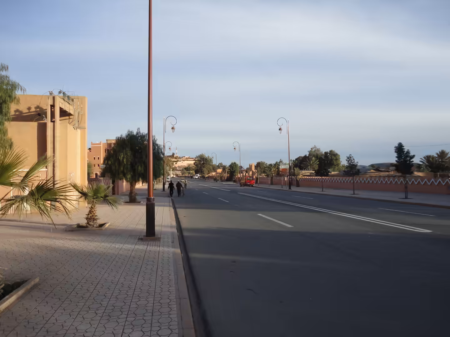 Wide boulevard in Ouarzazate with palm trees, decorative street lamps, pastel buildings, and a quiet evening atmosphere under a soft sky.