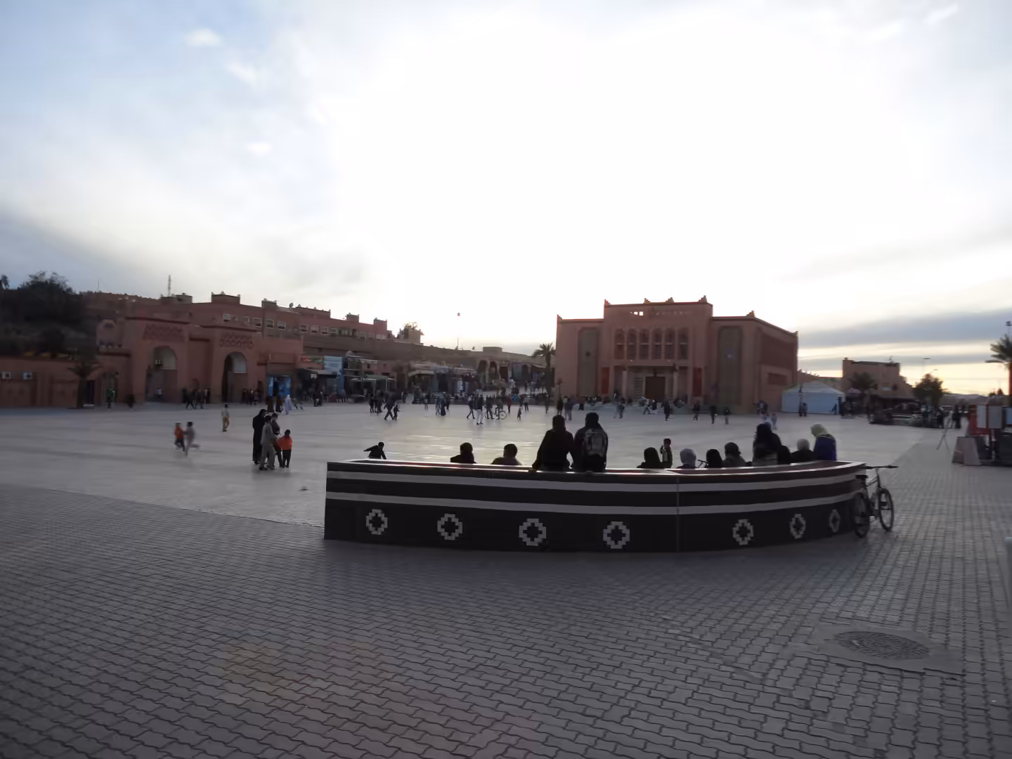 People gather in a wide public square at dusk in Ouarzazate, with pink-toned buildings and arched façades in the background.