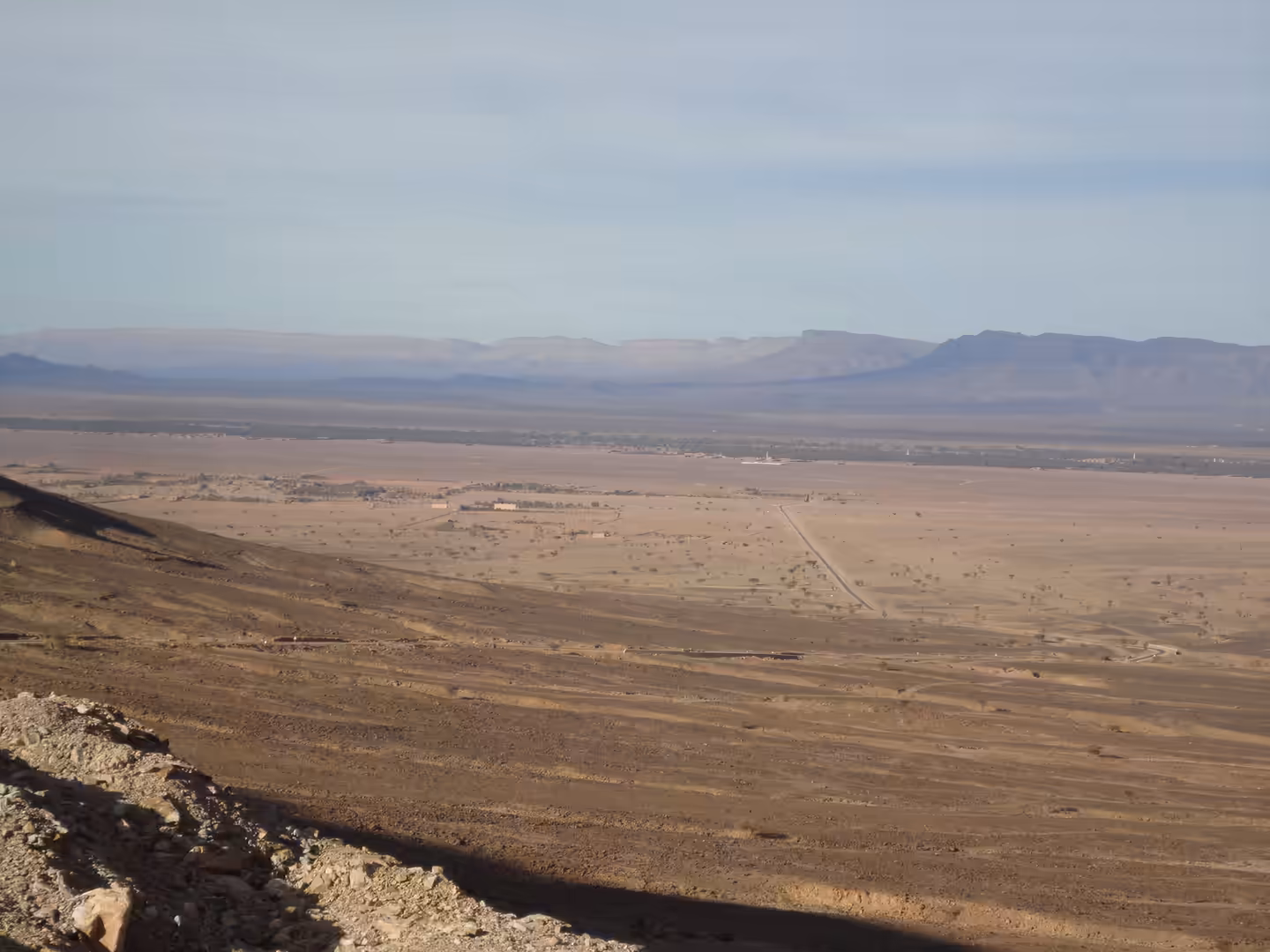 Wide arid valley with a straight road, sparse vegetation, and distant plateaus under a hazy sky.