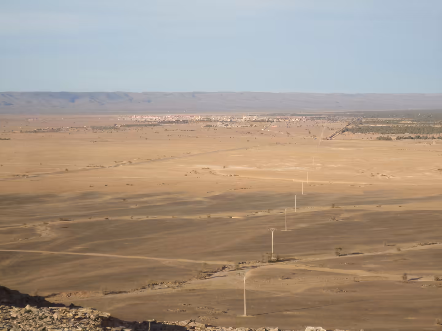 Wide desert plain with power lines leading toward a distant oasis town and palm groves under a pale blue sky.