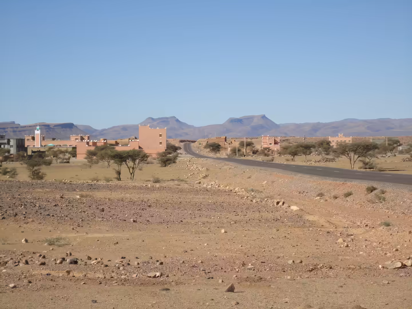 Desert road leading past a small village with adobe buildings and a mosque.