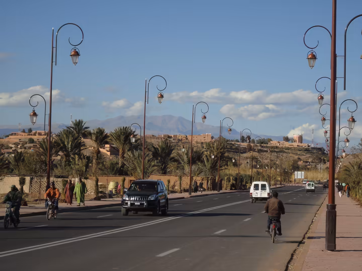Wide boulevard with decorative street lamps, palm trees, cyclists, cars, and distant mountains in Ouarzazate.
