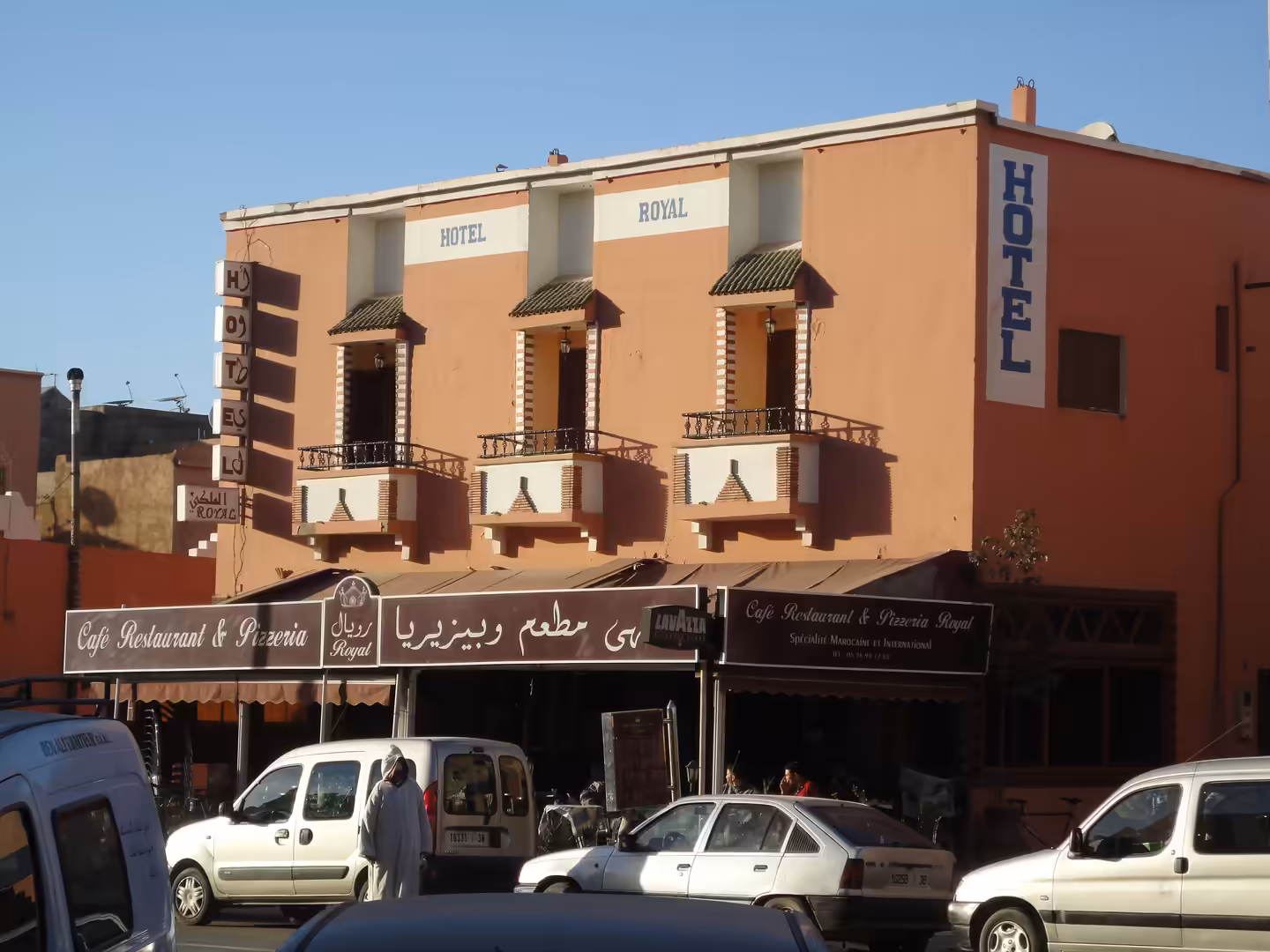 Peach-colored Hotel Royal with balconies above a café restaurant, cars parked along the street, and pedestrians in Ouarzazate.