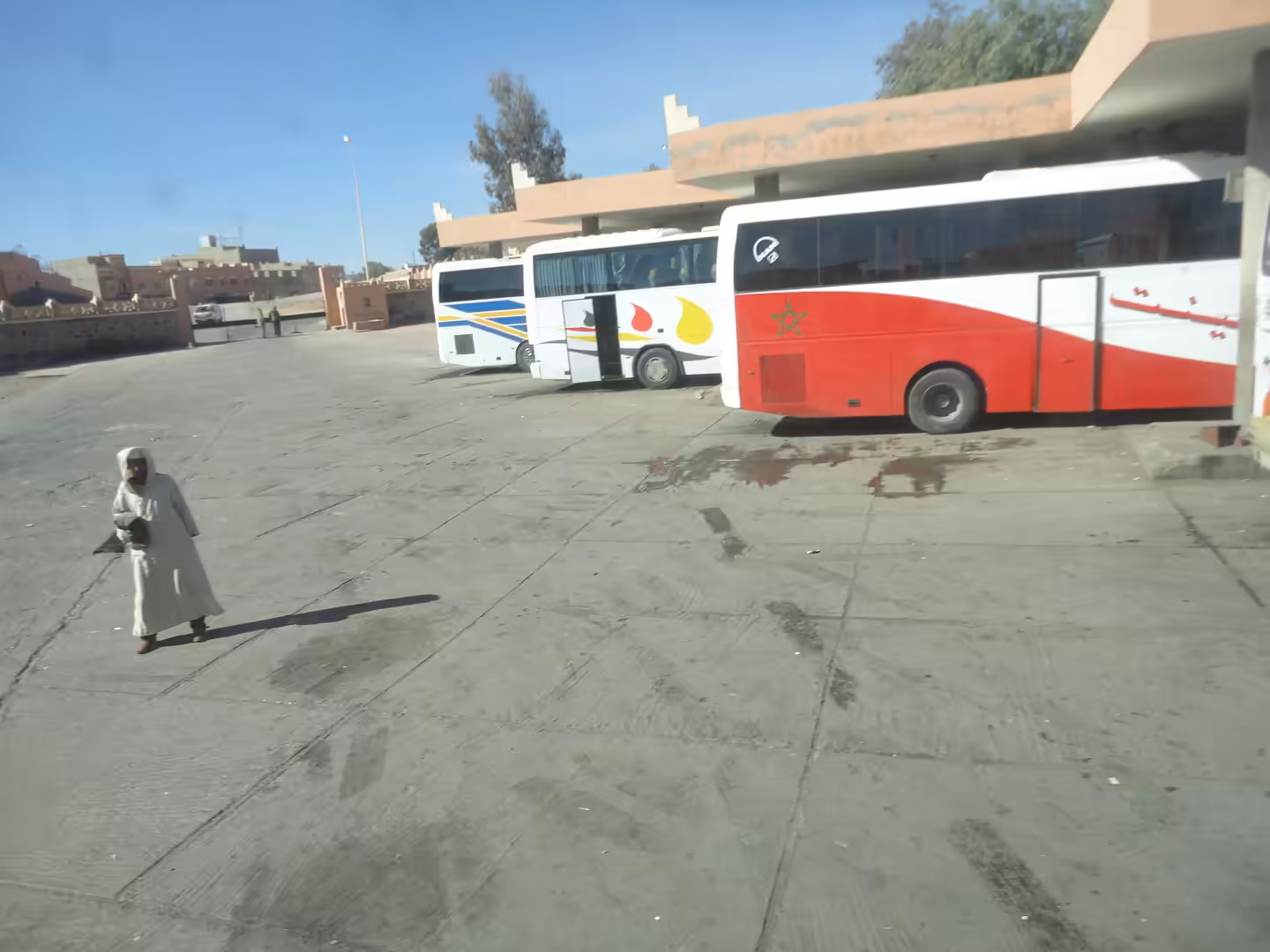 Intercity buses parked at a bus station with a pedestrian crossing the open concrete forecourt under clear skies.