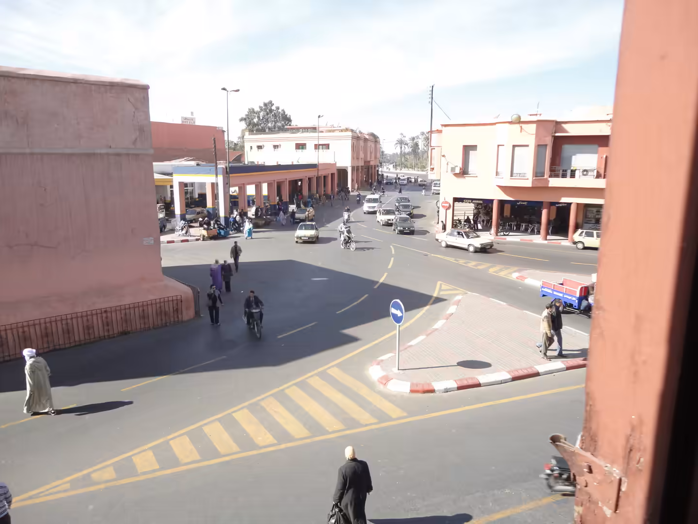 Busy urban intersection with pedestrians, motorbikes, cars, and pink-toned buildings at a city crossroads in Marrakech.
