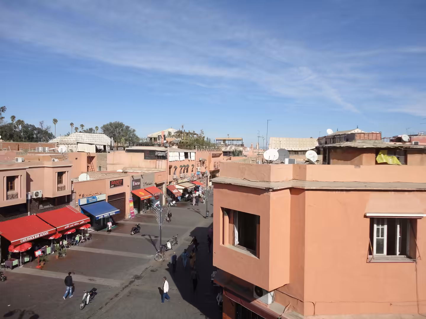 Elevated view over pink-toned buildings, cafés, shops, and pedestrians at a street junction in Marrakech.