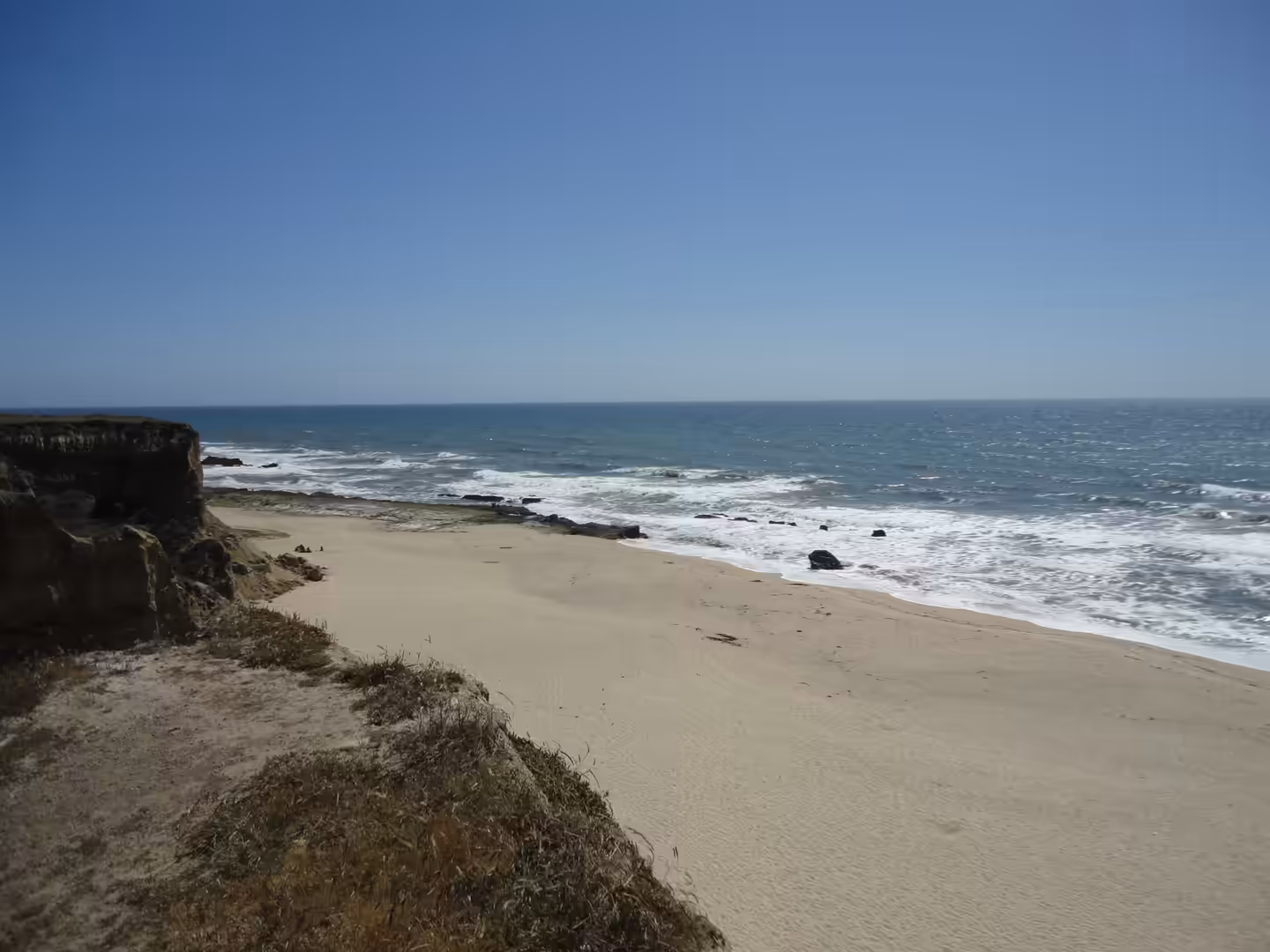 Sandy beach below low coastal cliffs with waves breaking along a rocky shoreline under a clear blue sky.