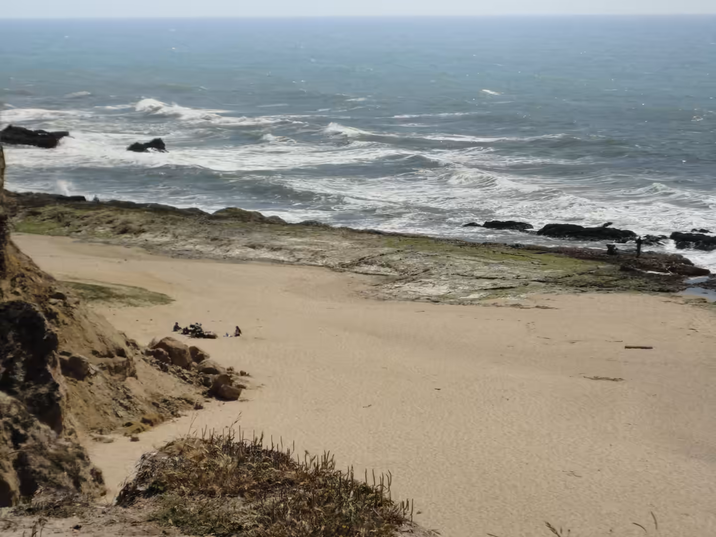 Small cove beach beneath sandy cliffs with tide pools and waves breaking along a rocky shoreline, viewed from above.