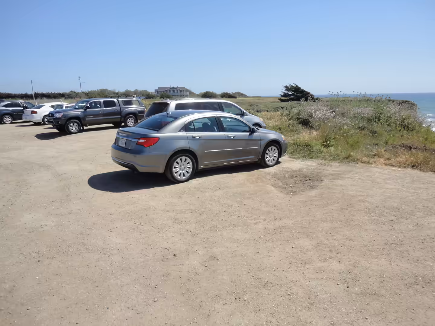A gray sedan parked in a dirt lot near coastal vegetation, with several other cars and the ocean visible in the background under a clear blue sky.