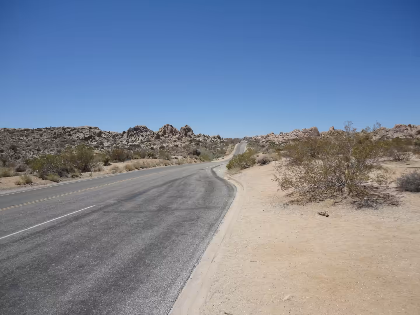A paved road curving through a rocky desert landscape with low shrubs under a clear blue sky