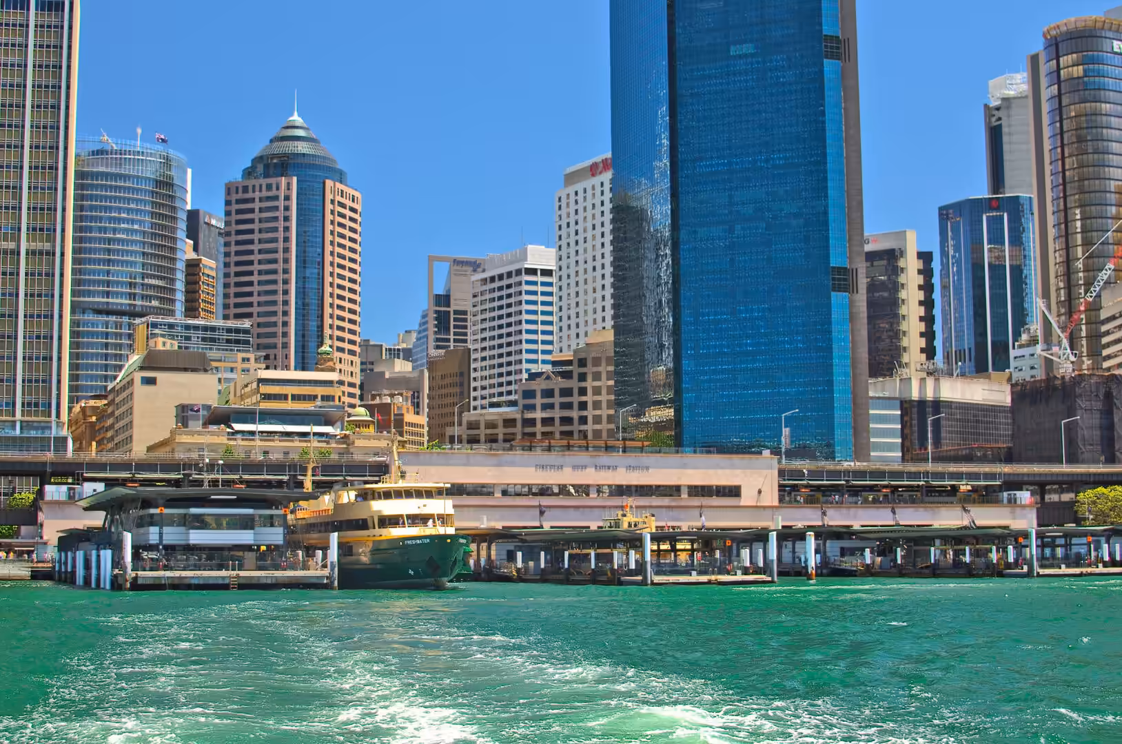 A green and gold ferry waits at a wharf beneath blue glass skyscrapers and a raised pedestrian walkway.