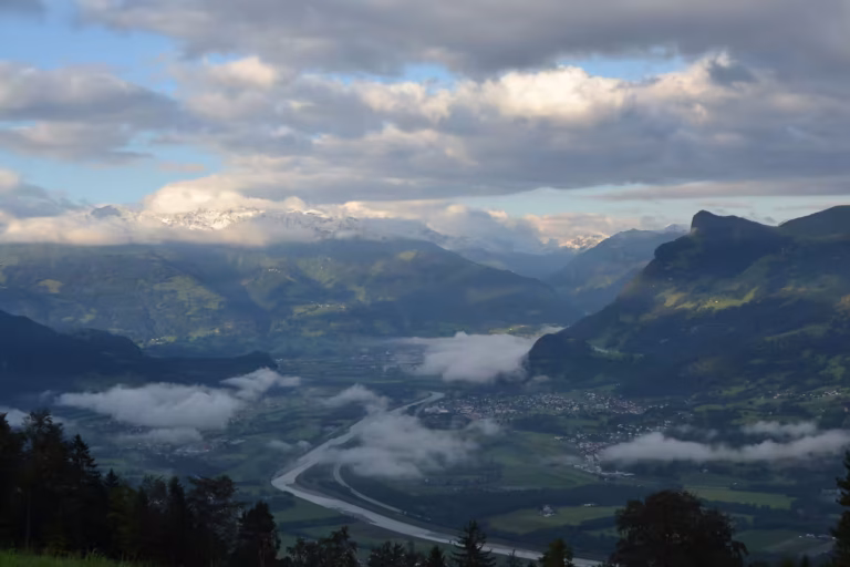 Aerial view over a broad alpine valley in Liechtenstein with a winding river, green meadows, banks of low cloud, and snow-capped peaks in the distance.