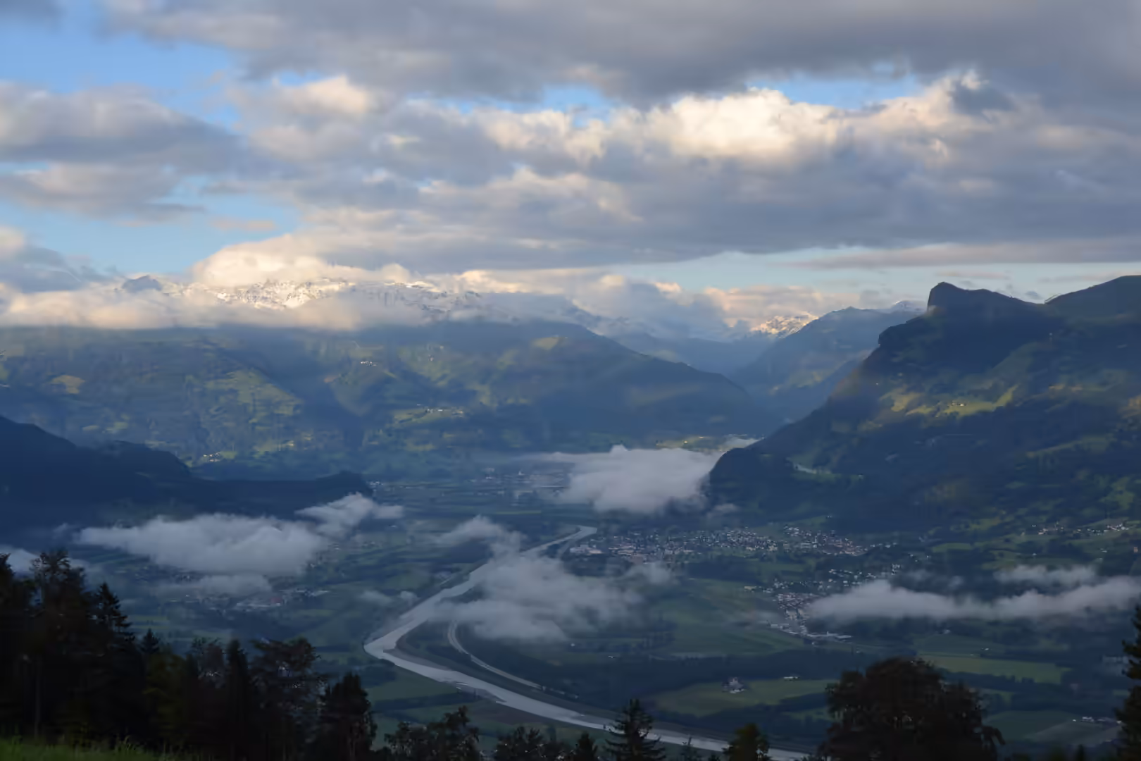 Aerial view over a broad alpine valley in Liechtenstein with a winding river, green meadows, banks of low cloud, and snow-capped peaks in the distance.