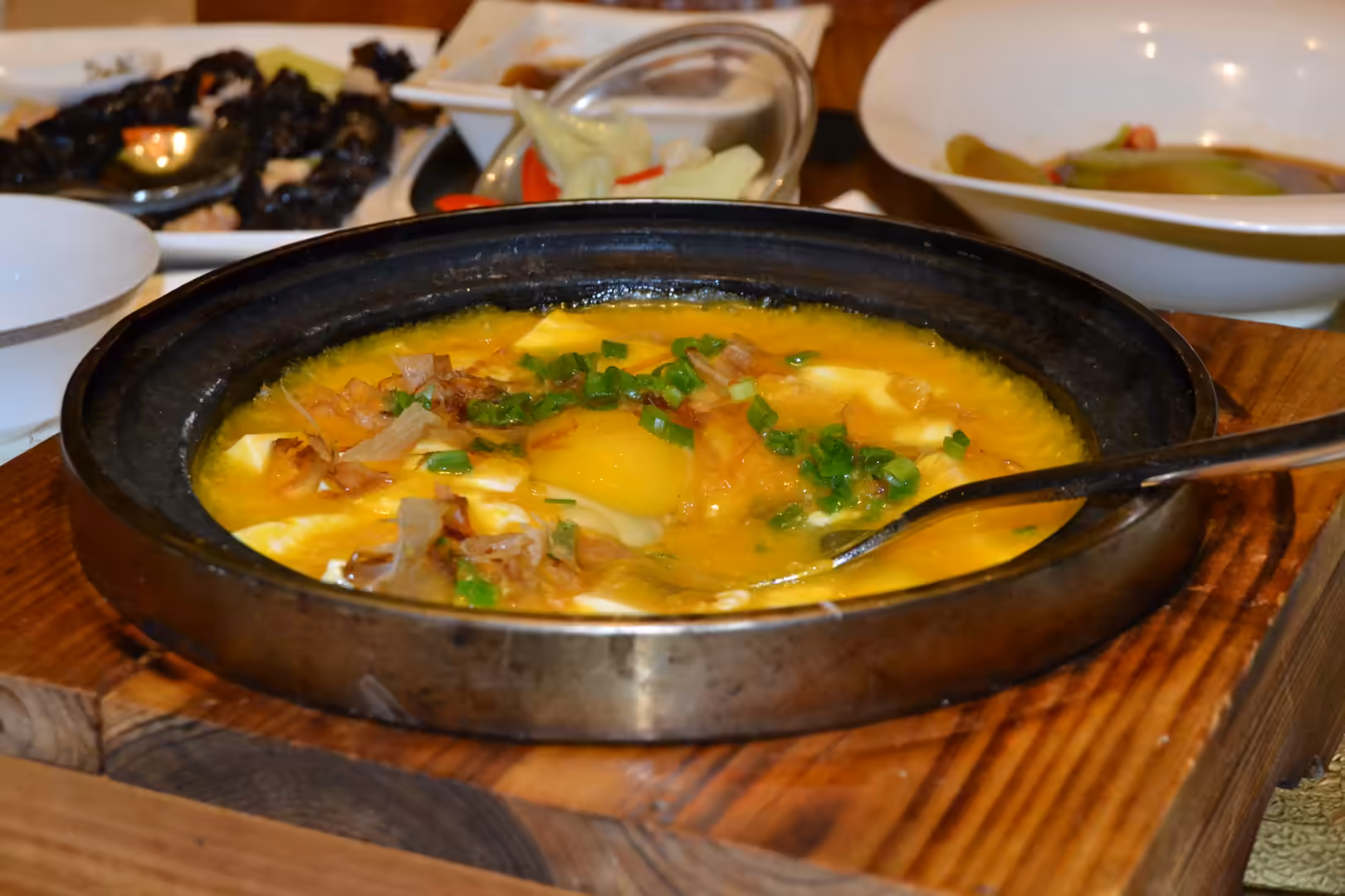 A hot Chinese egg and tofu dish topped with chopped scallions and fried shallots, served in a black pan on a wooden board.