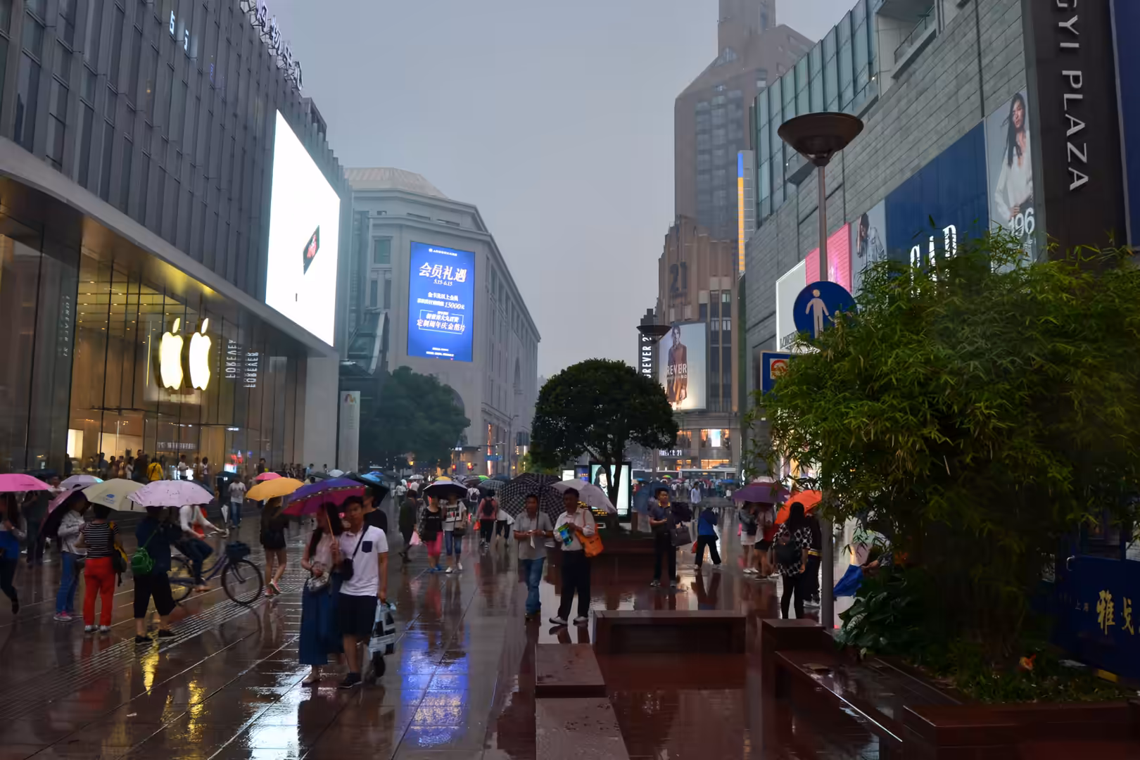 Pedestrians carrying colorful umbrellas walk along a rain-soaked shopping street lined with illuminated stores and large digital advertisements in Shanghai.