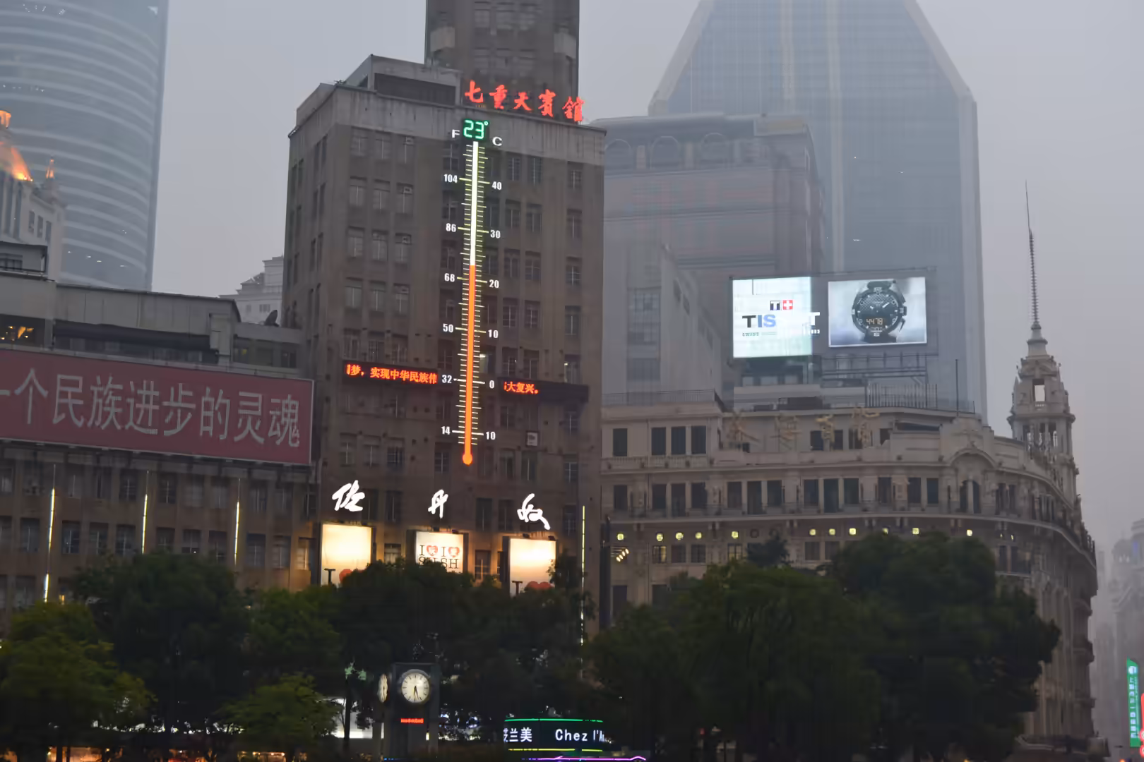 Historic and modern buildings rise through dense haze in central Shanghai, with illuminated signs, billboards, and a large vertical temperature display.