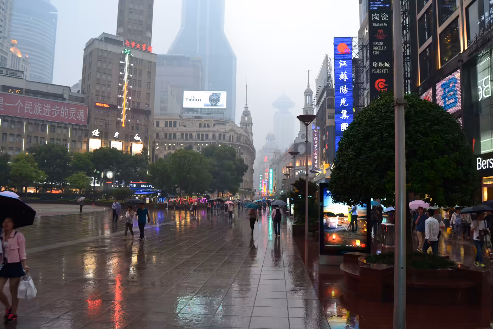 Pedestrians with umbrellas walk along a rain-soaked Nanjing Road, surrounded by illuminated signs, shops, and hazy high-rise buildings in Shanghai.