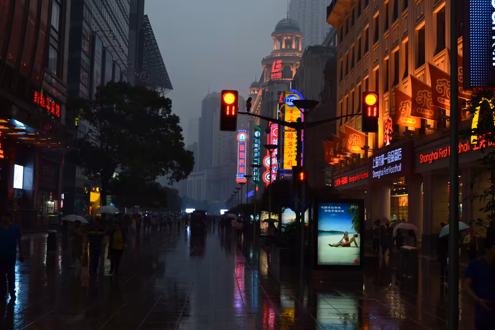 Pedestrians with umbrellas walk along rain-soaked Nanjing Road beneath glowing neon signs and red traffic lights in Shanghai.