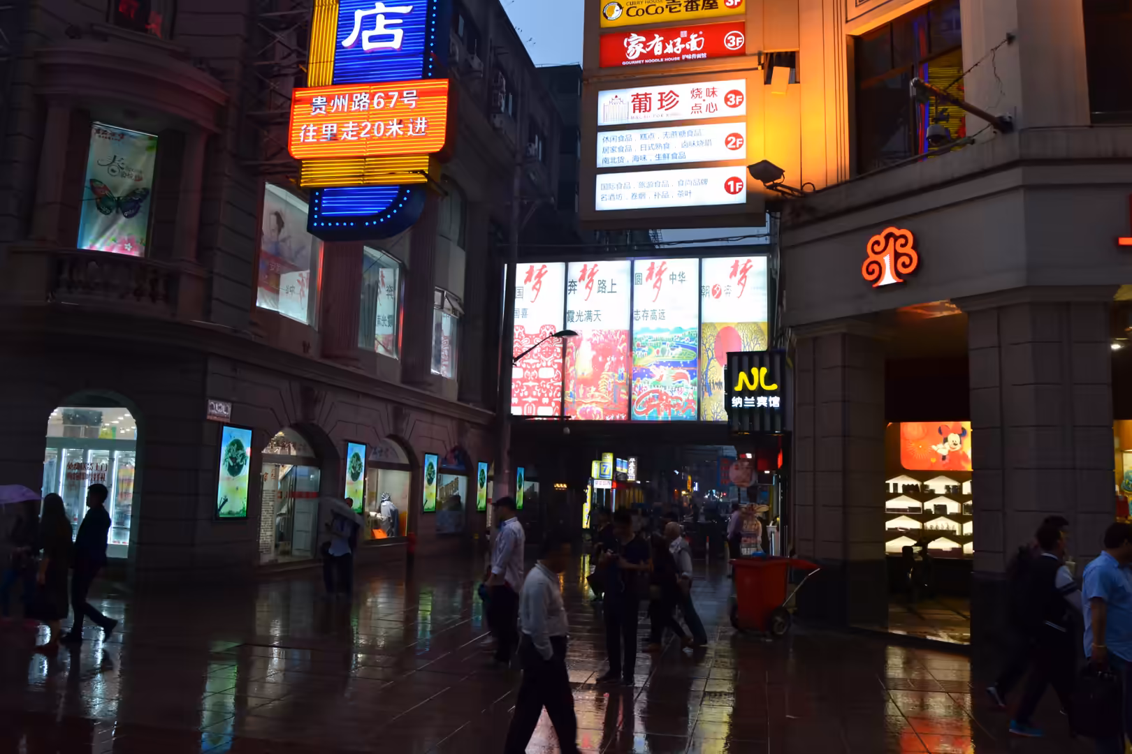 Pedestrians walk through a rain-soaked commercial passage illuminated by colorful Chinese signs and storefront displays in Shanghai.