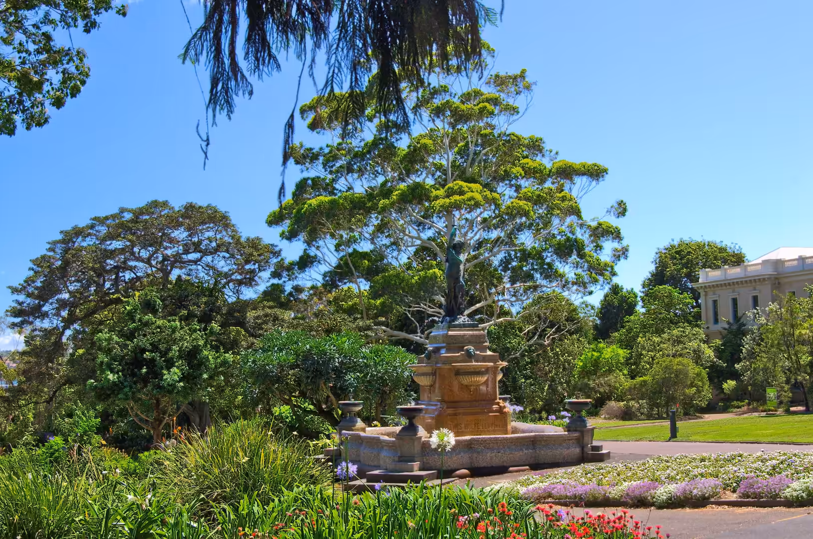 A dark statue tops a stone fountain ringed by flower beds and lawns.