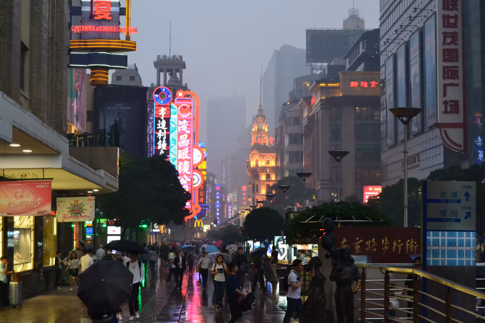 Crowds carrying umbrellas walk along rain-soaked Nanjing Road beneath glowing neon signs and hazy high-rise buildings in Shanghai.