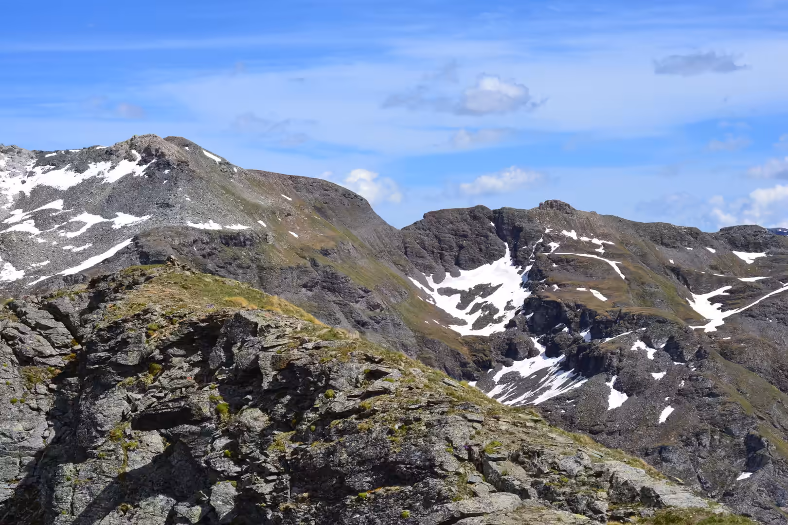 Craggy ridges of grey rock hold patches of snow, with green tufts on the lower ledges under a blue sky.