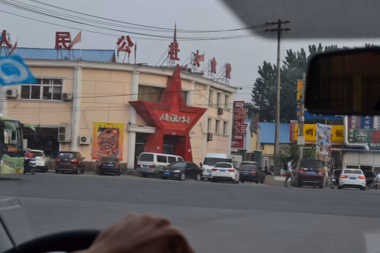 Street scene in Beijing, China, with a public dining complex marked by a large red star entrance, Chinese signage, parked cars, buses, and overhead utility lines.
