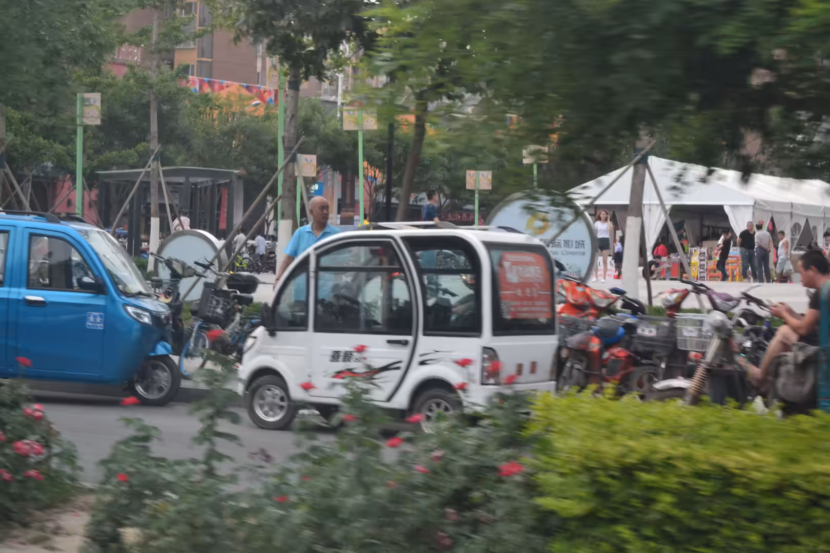 Small electric vehicles and parked bicycles in a leafy residential area of Beijing, China, with pedestrians and a temporary community tent in the background.
