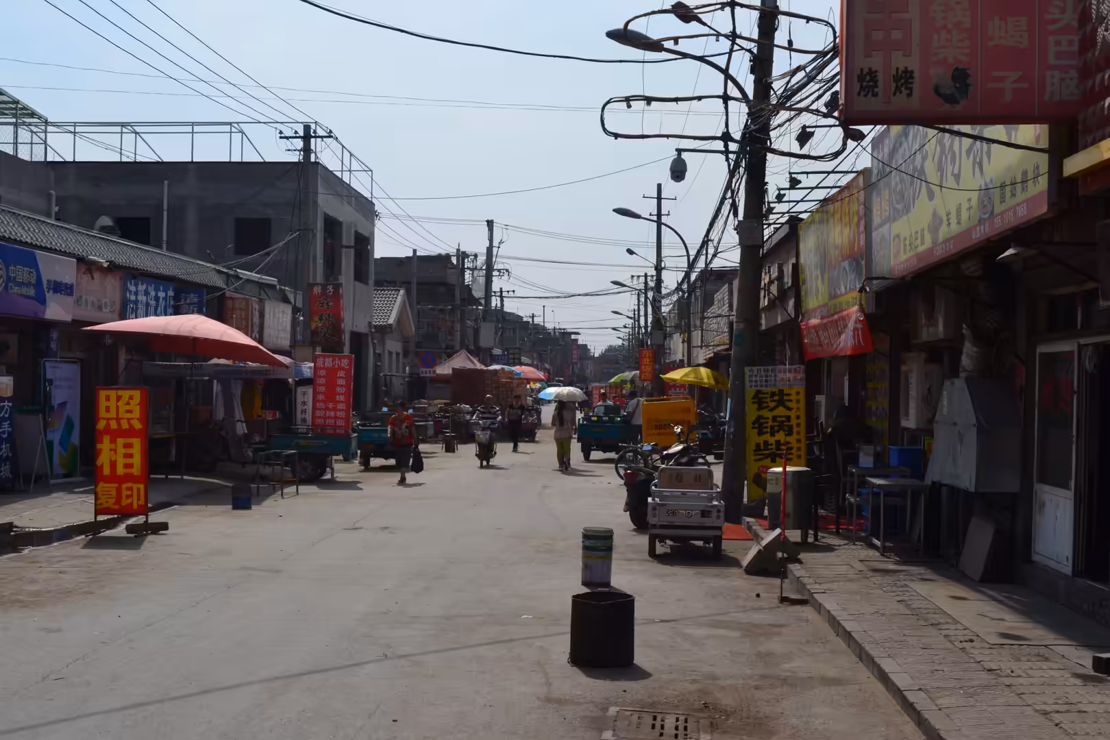 Narrow neighborhood street in Beijing, China, lined with small shops, food stalls, scooters, three-wheeled vehicles, pedestrians, colorful signs, and dense overhead wiring.