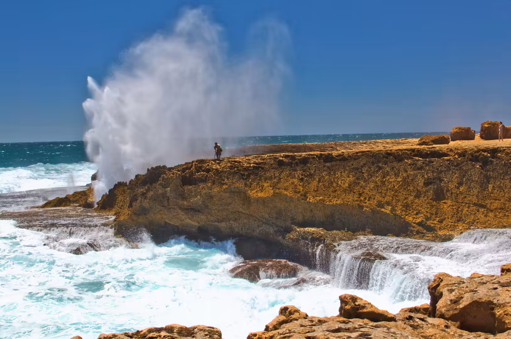 A lone figure stands on the rock edge while ocean spray erupts behind them.