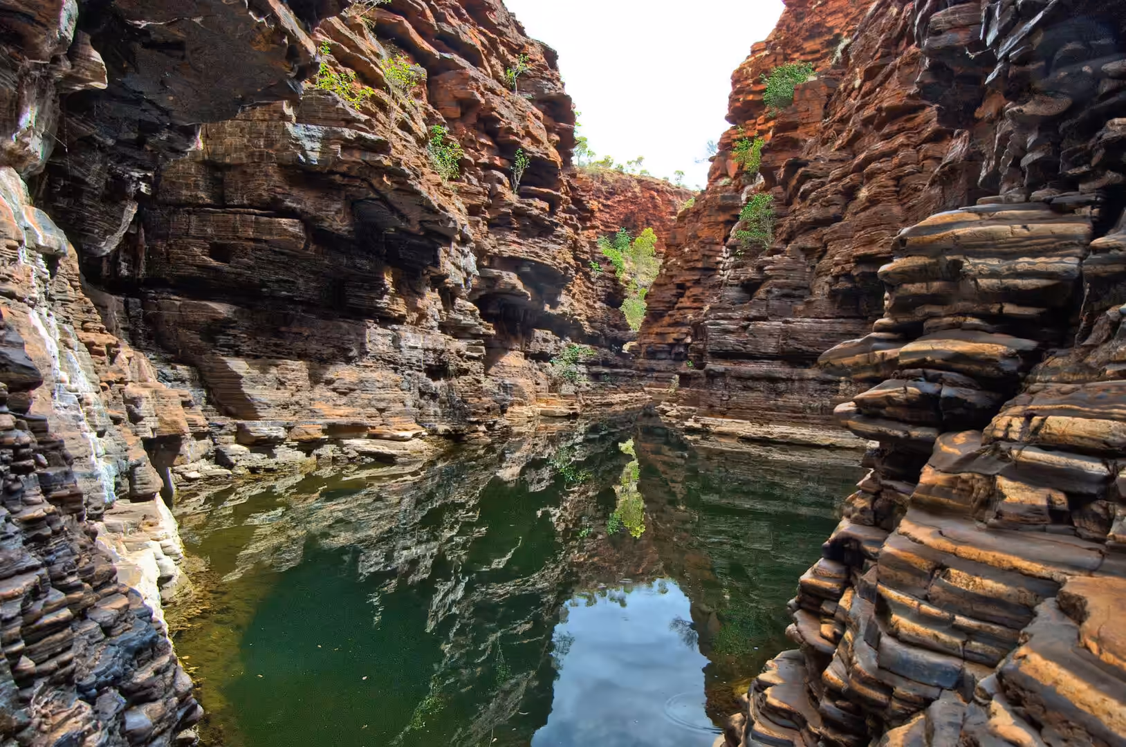 A dark green waterhole sits between high, banded red rock walls that mirror in the surface.