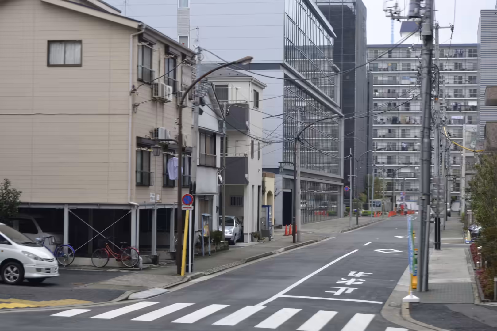 A quiet residential street in Tokyo lined with low houses and apartment blocks, with a zebra crossing, parked cars and overhead power lines.