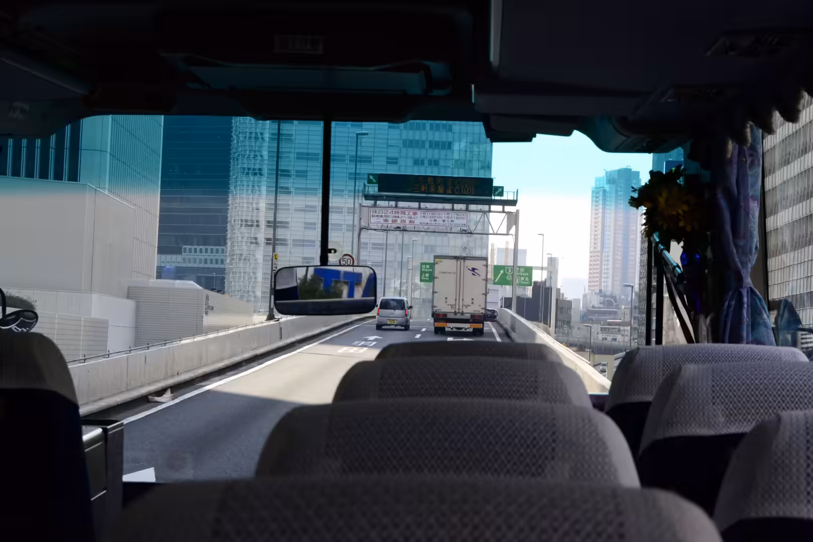 From inside a coach on an elevated expressway, a white truck drives beneath a green direction sign toward glass skyscrapers.