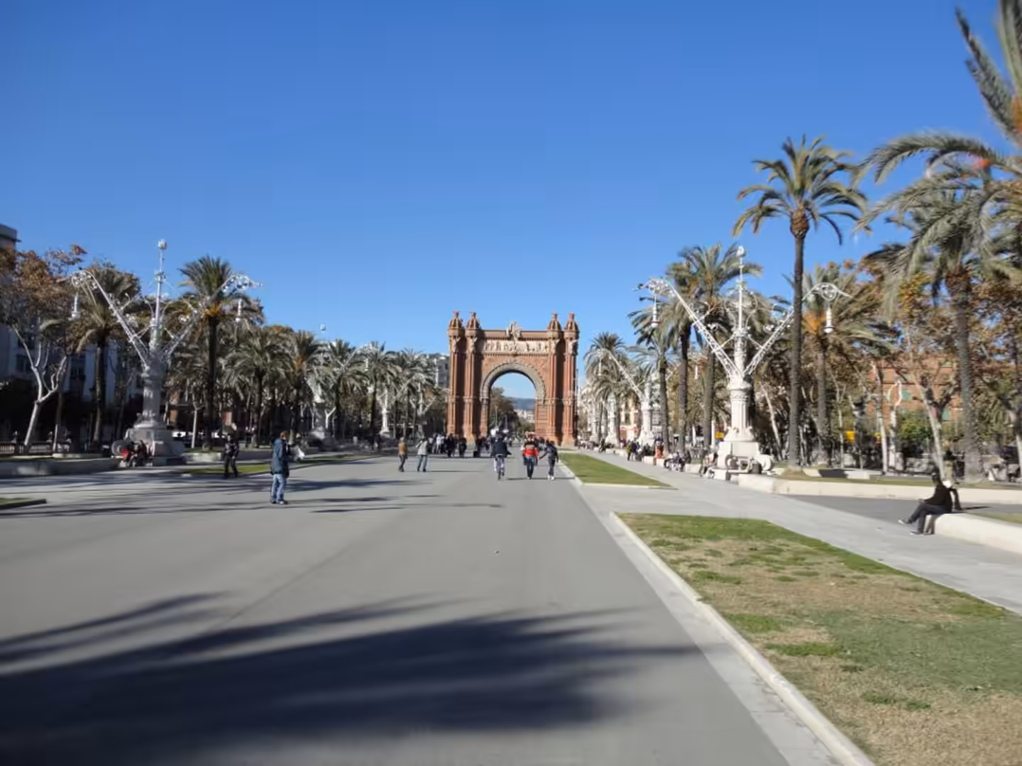 Wide pedestrian avenue lined with palm trees leading to the Arc de Triomf in Barcelona, with people walking and cycling on a sunny day.