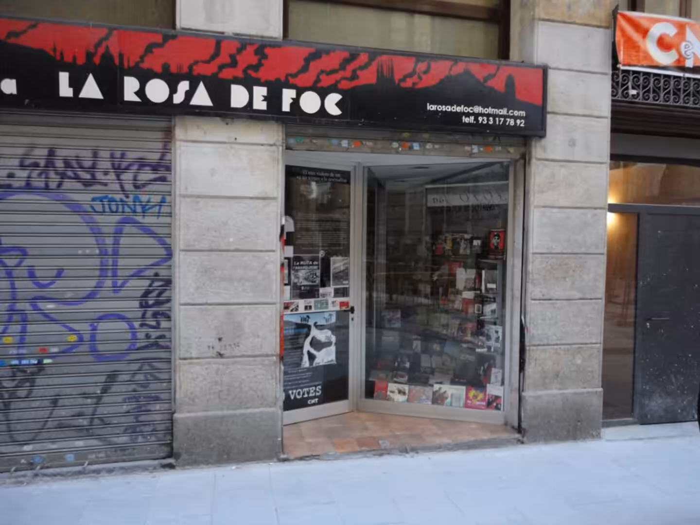 Small bookstore entrance in Barcelona with the sign “La Rosa de Foc,” books visible through the glass door, and graffiti on a closed metal shutter beside it.