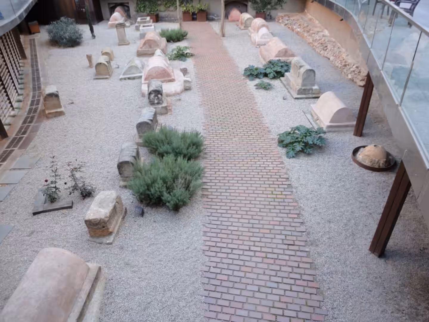Indoor courtyard with a central brick walkway surrounded by gravel, ancient stone fragments, carved blocks, and small plants.
