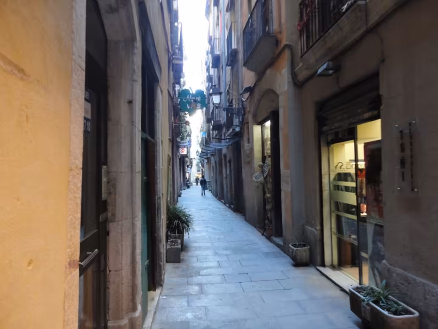 Narrow old-city alley paved with stone, lined by tall buildings, small shop entrances, balconies, and a few pedestrians in the distance.