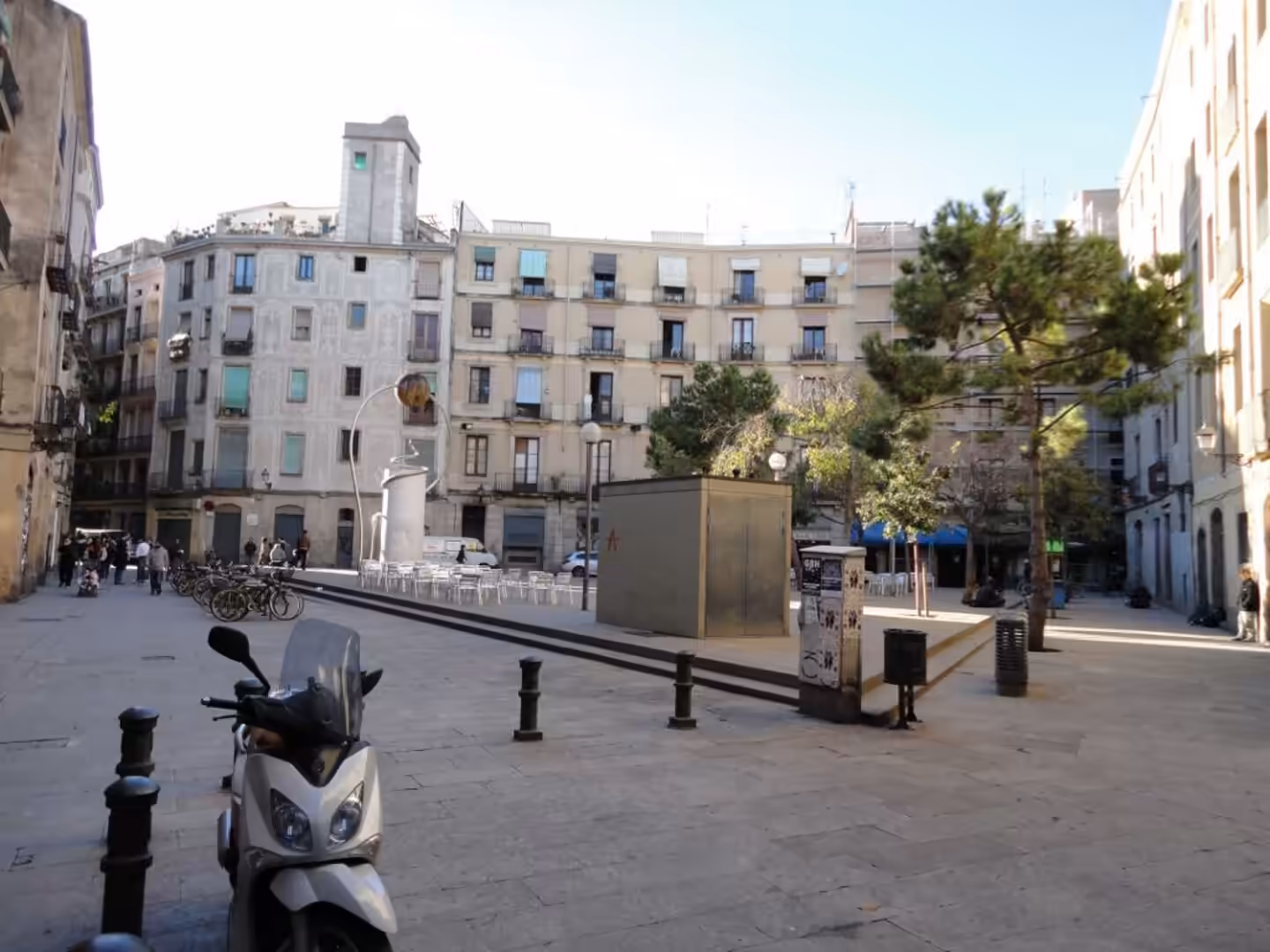Open city square in Barcelona with surrounding apartment buildings, a scooter in the foreground, and outdoor café tables.
