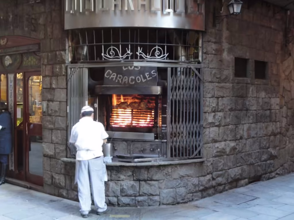Chef in white uniform cooking over a wood-fired grill behind iron bars in a stone building in Barcelona.