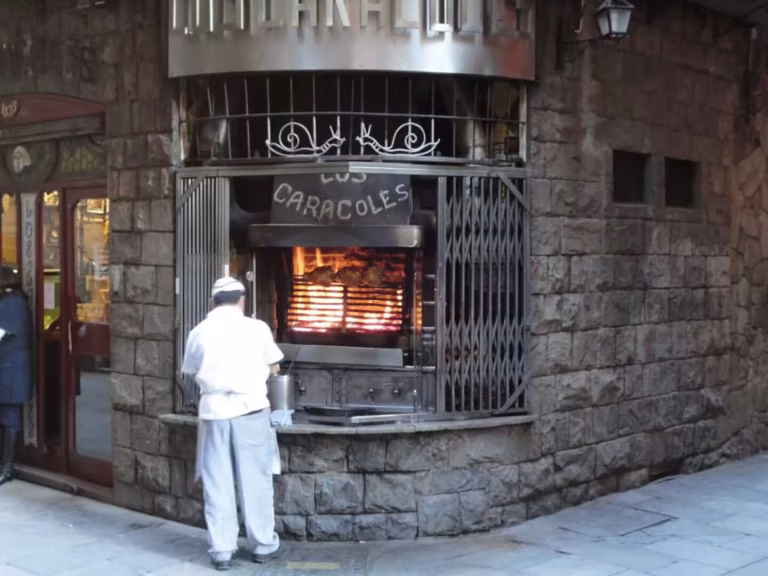Chef in white uniform cooking over a wood-fired grill behind iron bars in a stone building in Barcelona.