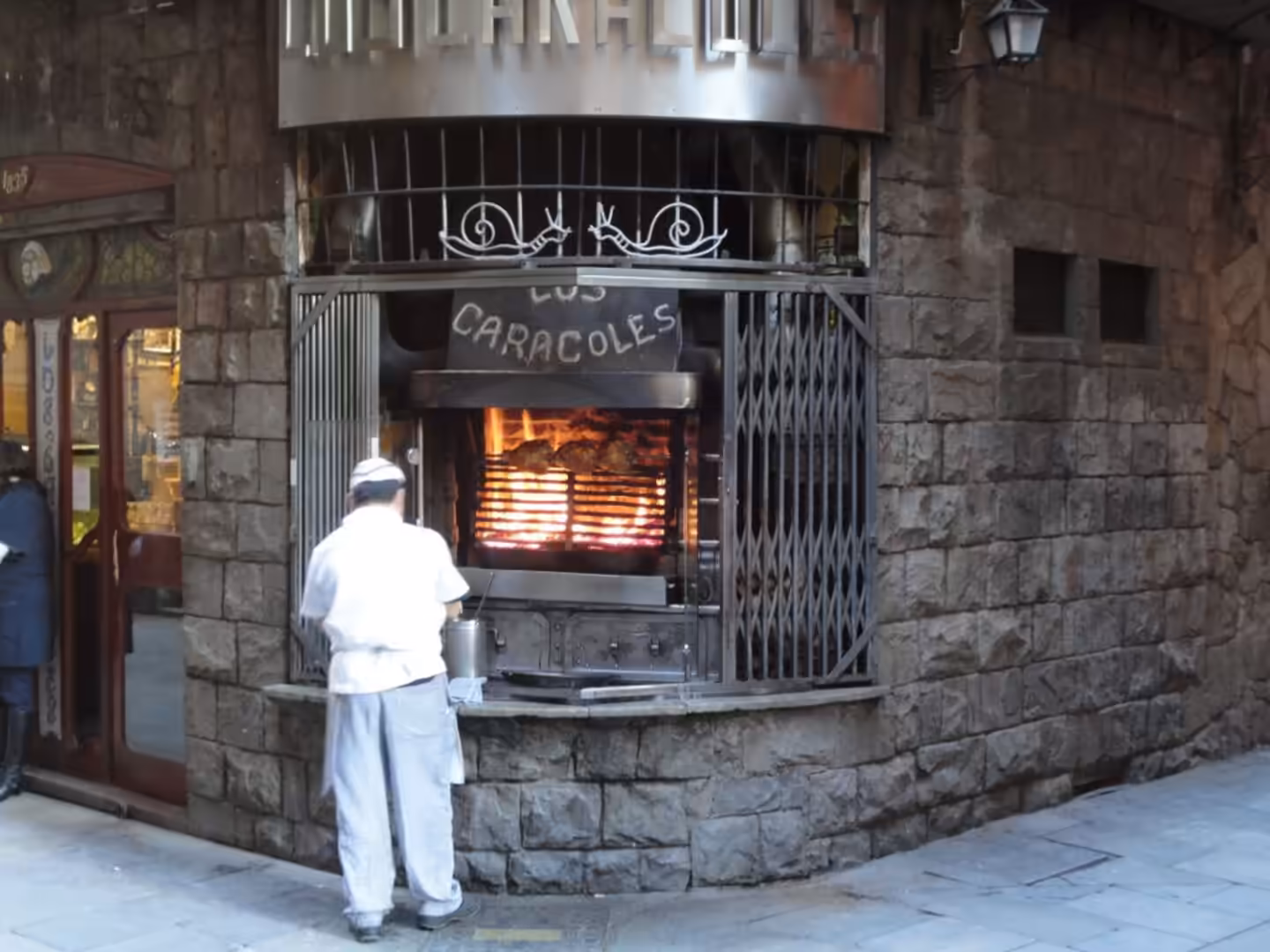 Chef in white uniform cooking over a wood-fired grill behind iron bars in a stone building in Barcelona.