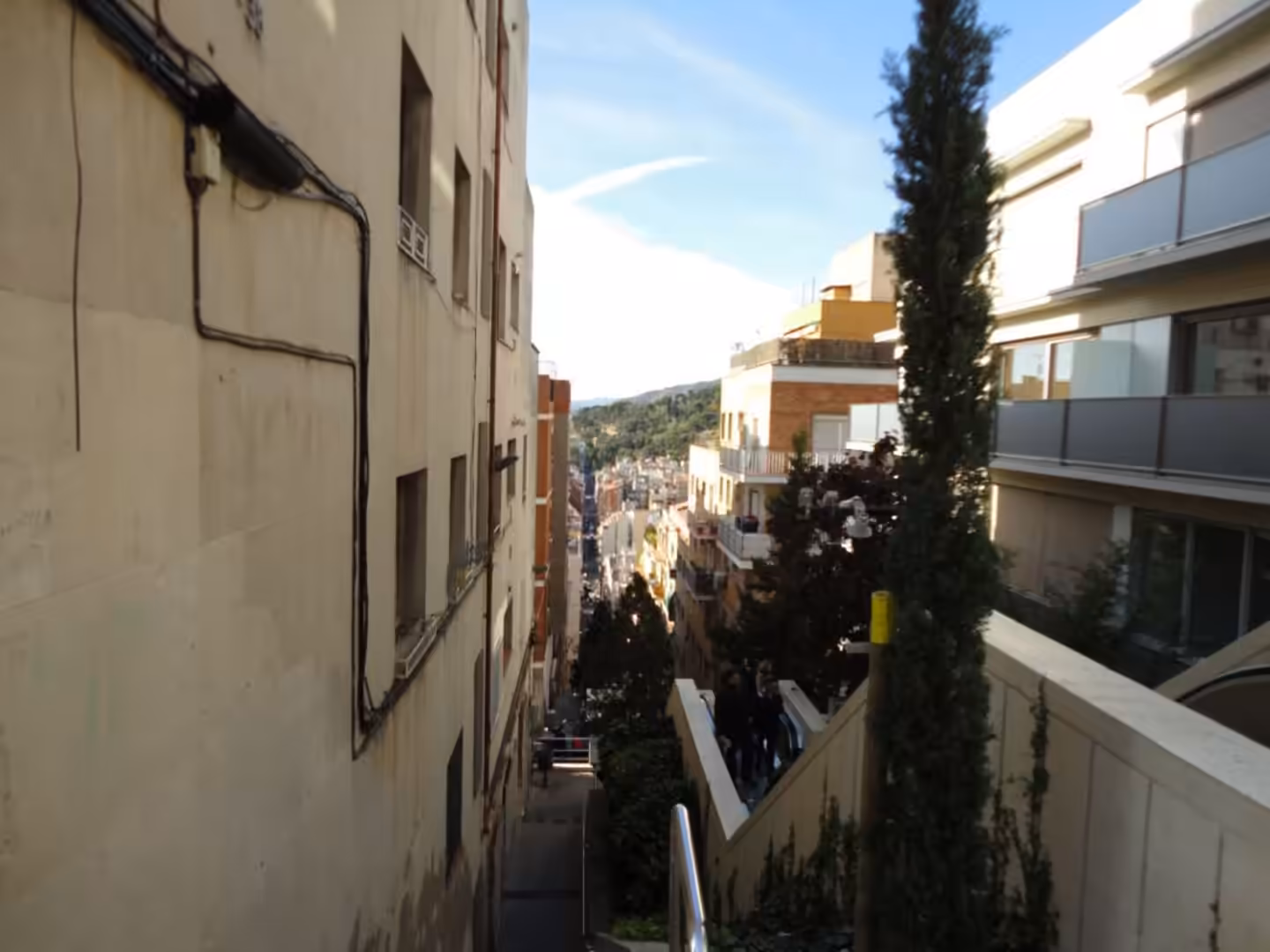 Steep outdoor staircase between apartment buildings in Barcelona, looking down toward the city with trees and hills in the background.
