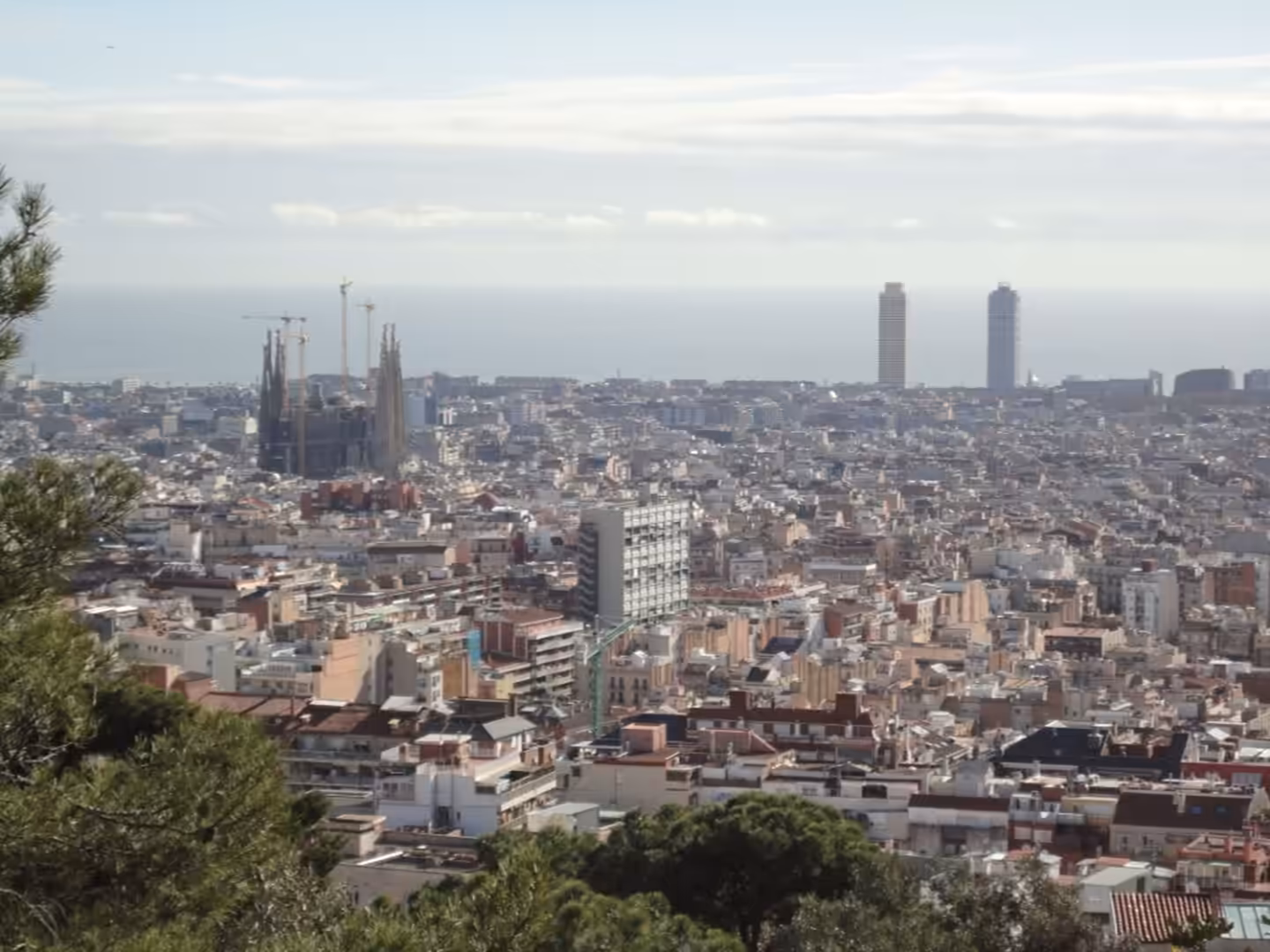 Panoramic view of Barcelona from an elevated park, showing the Sagrada Família, dense urban rooftops, and the Mediterranean Sea in the distance.