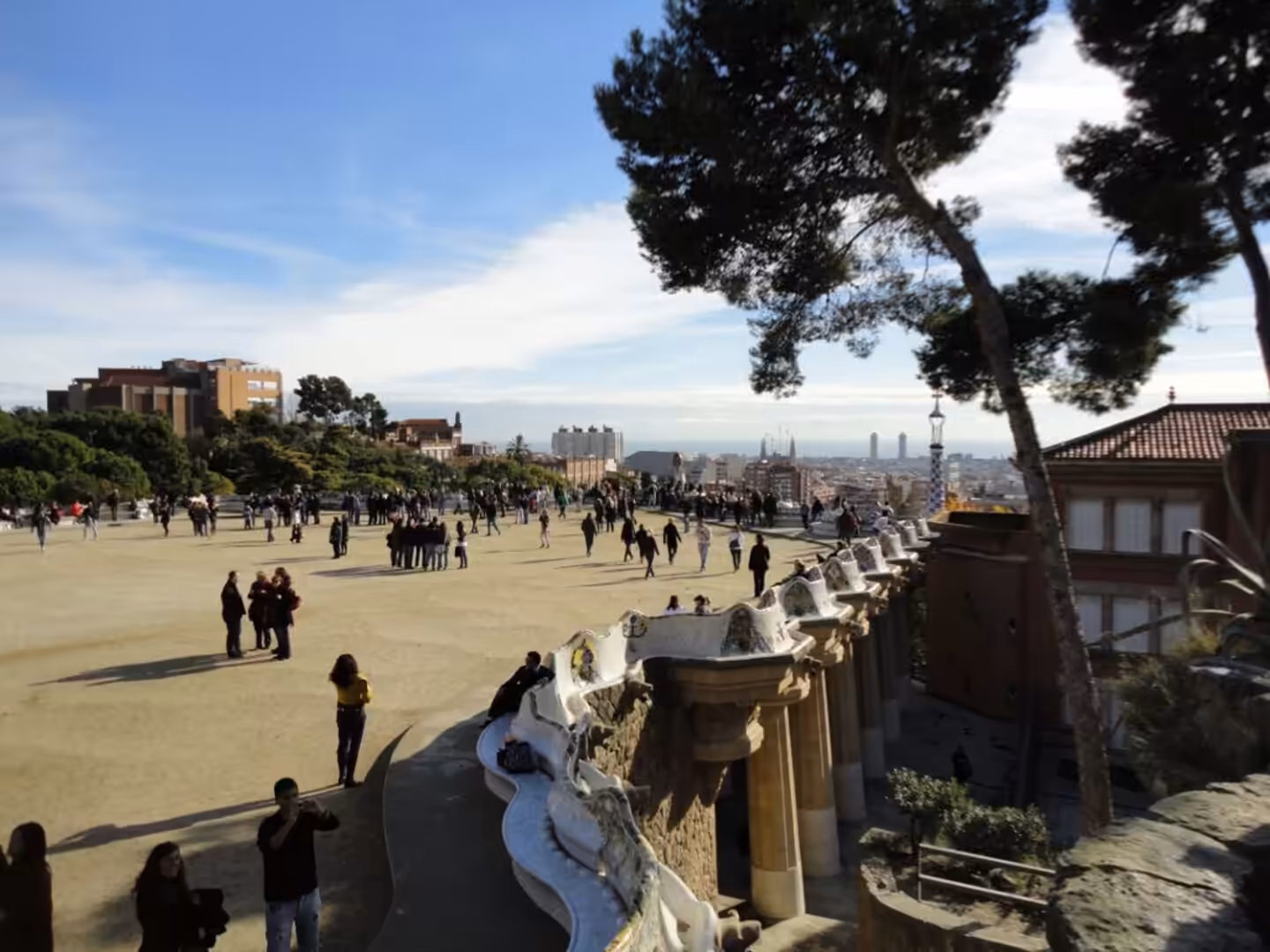Crowds walking and standing on the terrace at Park Güell in Barcelona, with mosaic benches, trees, and the city skyline in the distance.
