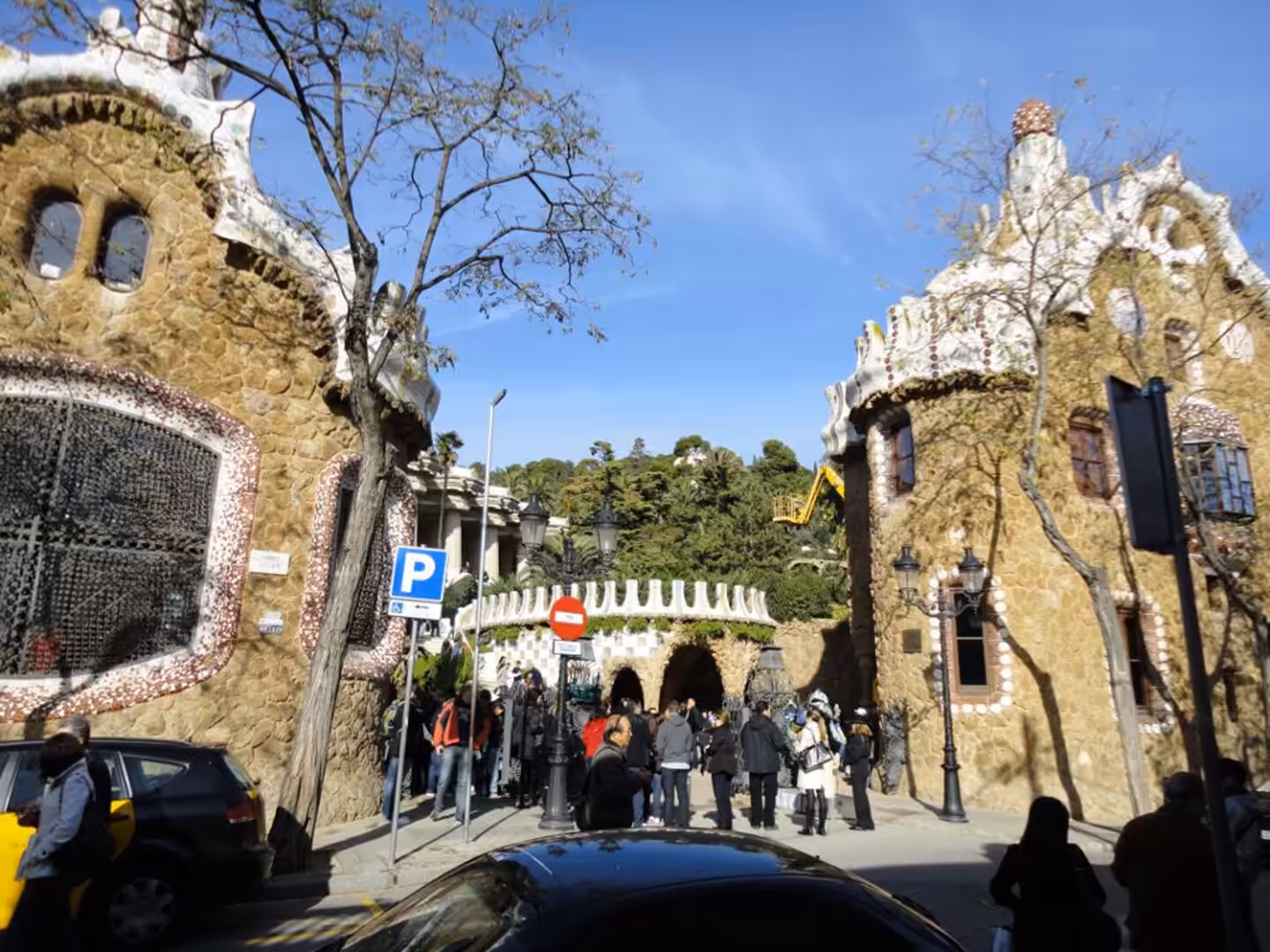 Crowds entering Park Güell through Gaudí-designed stone buildings with mosaic roofs under a clear blue sky in Barcelona.