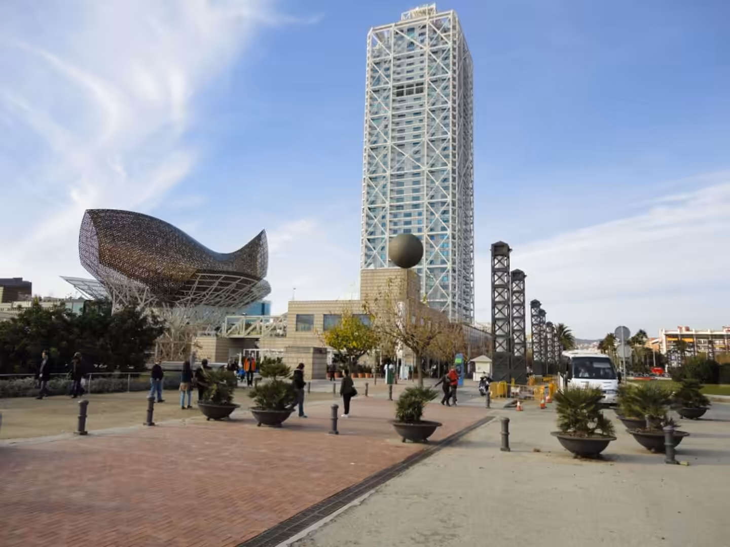 Open plaza with people walking, a tall glass-and-steel skyscraper, and a curved metallic building at Port Olímpic in Barcelona.